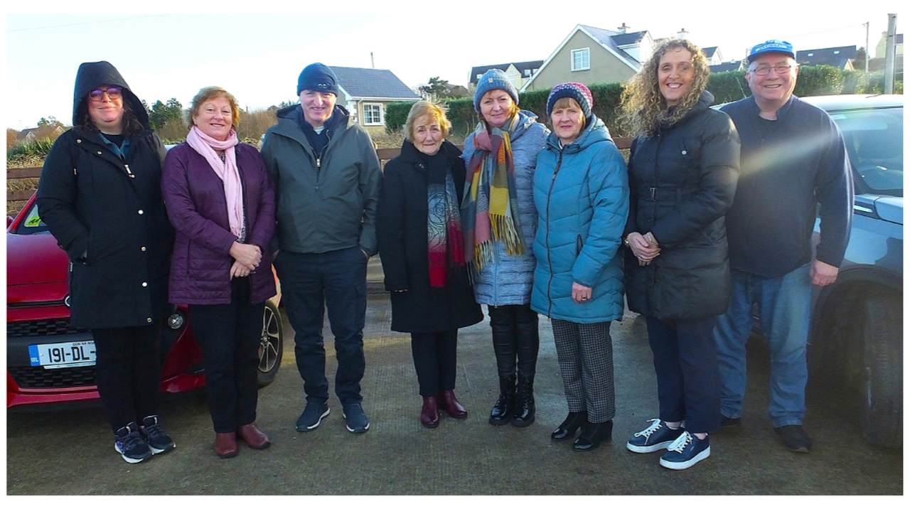 A group of eight people standing outdoors, dressed in winter clothing, smiling at the camera with houses and cars in the background.